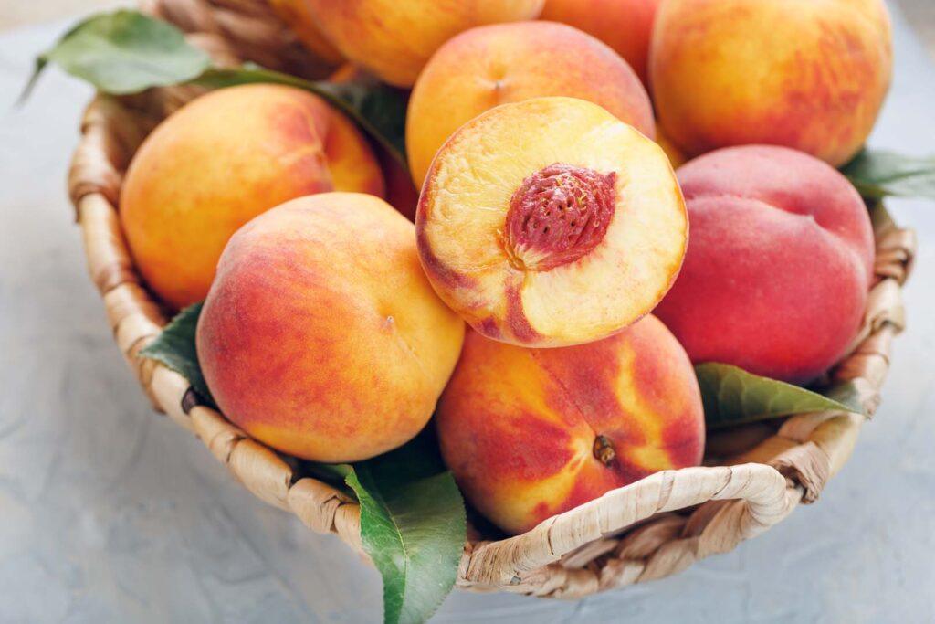 Ripe peaches in a wicker basket on a stone gray table with a juicy peach slice with a stone pit. Top view, free space.